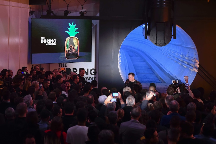 Elon Musk walks into awaiting crowd from the right with a blue tunnel behind him, as attendees at unveiling event take pictures.