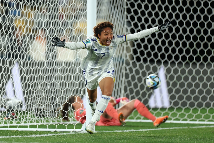 A women's footballer from the Philippines grins as she runs away with arms spread as the ball lies in the net with the keeper.