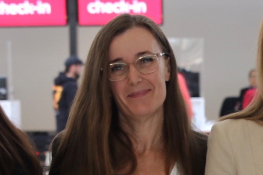 A close-up, cropped image of Renee Freeman in an airport terminal with a red check in sign behind her.