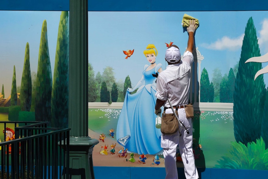 A man wearing white cleaning overalls scrubs a wall with a cloth. A bright coloured cartoon picture of Cinderella is on the wall