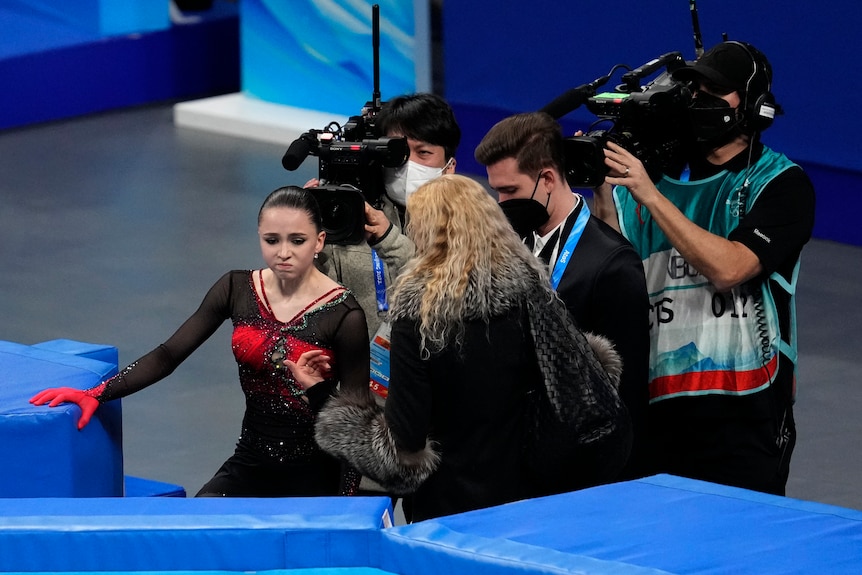 A sad-looking figure skater holds on to a barrier at the Winter Olympics as her coach talks to her and cameras film them.  