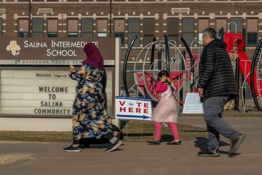 A family including a woman in a headscarf walk past a school with a sign that says vote here.