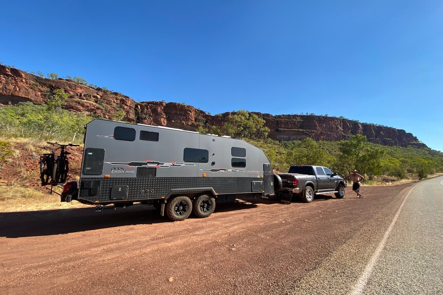 A large caravan and ute on a remote outback road with a man standing at the front