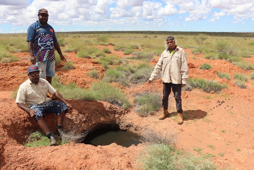 Three men stand outside in the desert pointing at a small waterhole