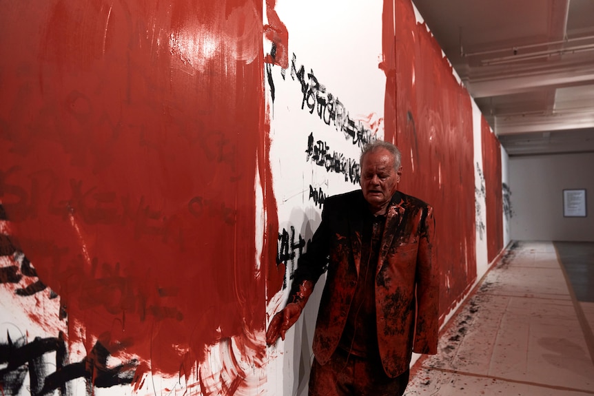 Mike Parr performing in a gallery space with his eyes closed, covered in red paint beside a wall covered in black and red paint.