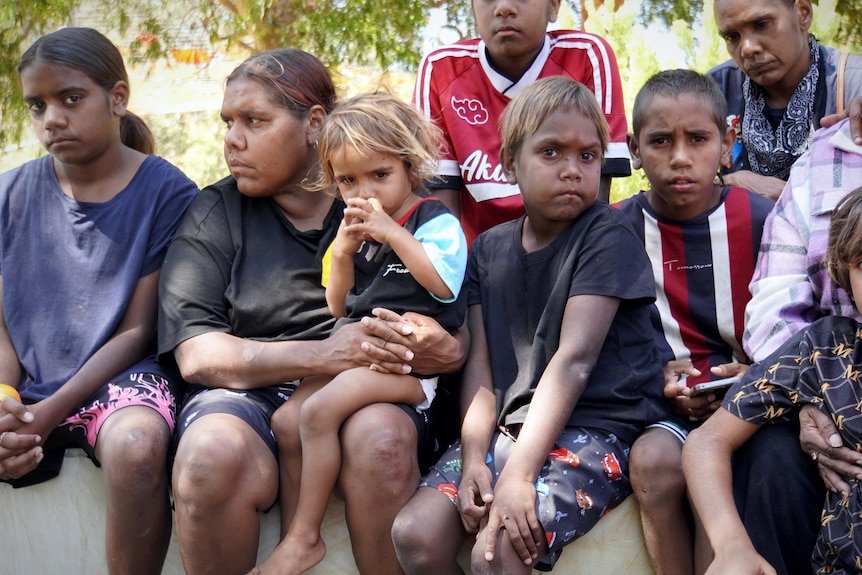A group of Indigenous family members gathered in a park looking sombre and embracing each other