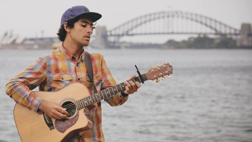 A man playing his guitar, with a view of a large bridge in the background.