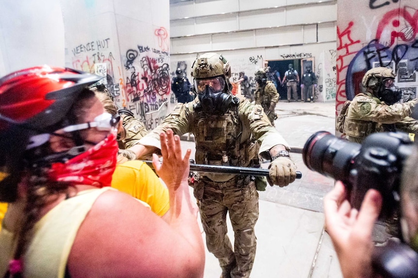 A federal officer uses a baton to push back demonstrators from a building covered in graffiti.