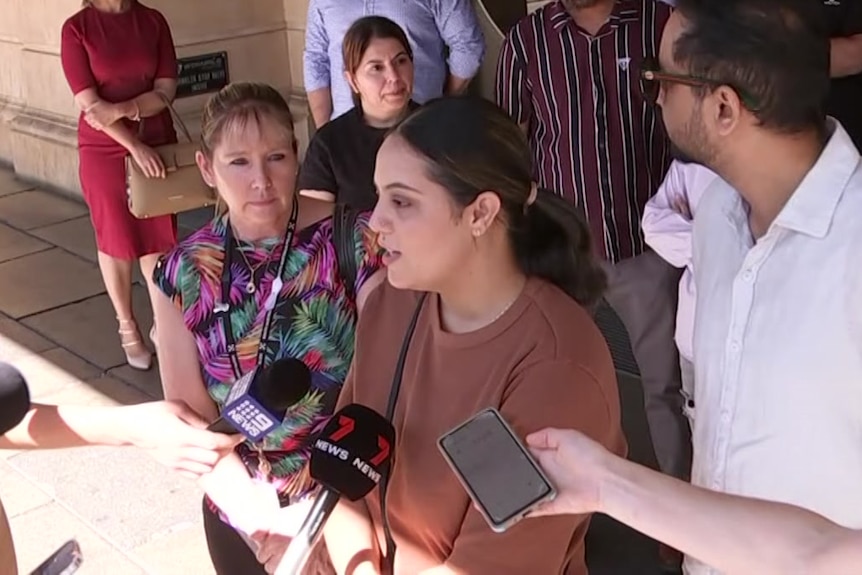A woman with black hair wearing a brown top speaks to reporters