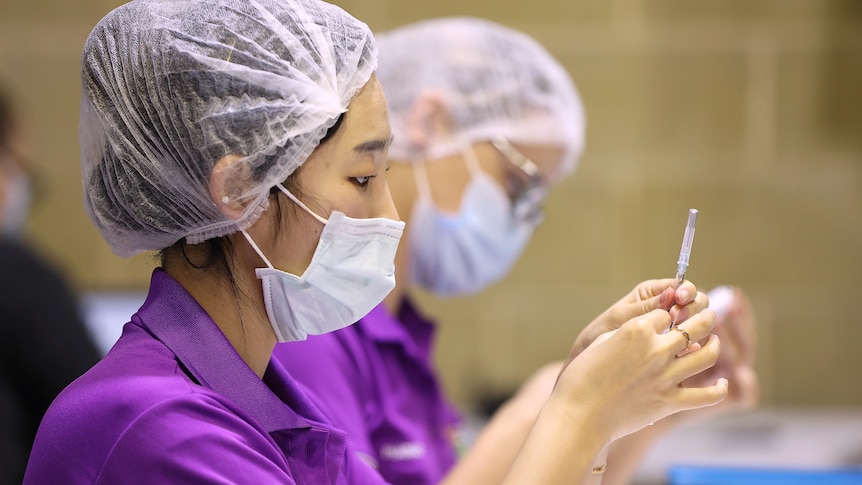 Woman dressed in purple t-shirt, wearing a hair net and face mask draws up vaccine into a syringe.