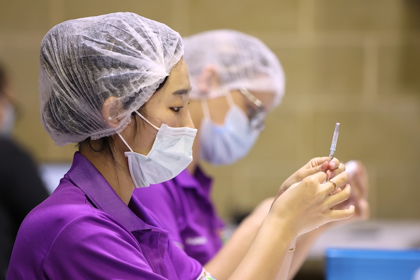 Woman dressed in purple t-shirt, wearing a hair net and face mask draws up vaccine into a syringe.