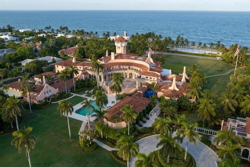 An aerial photograph shows a sprawling mix of pink and beige buildings and palm trees on a beachfront property.