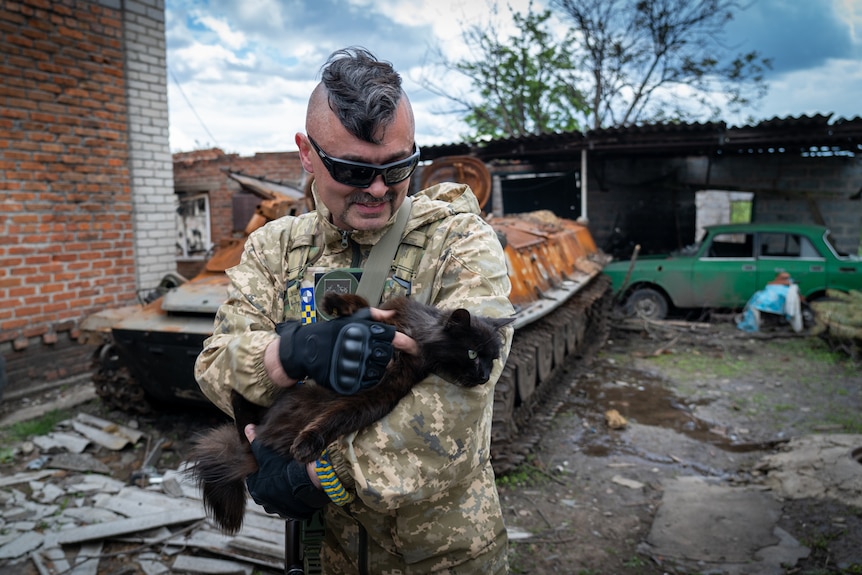 A soldier in army fatigues with a thick mohawk and dark shades smiles adoringly at a black cat in his arms