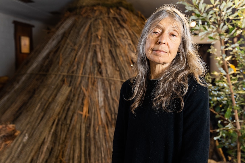 A 67-year-old woman stands in front of an art installation of a bark hut set inside a shopfront