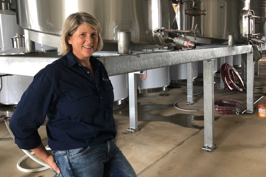 A lady wine maker stands in front of large wine tanks used for making wine.