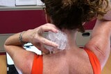 A woman with her back to the camera holds ice to the back of her neck as she sits at a desk