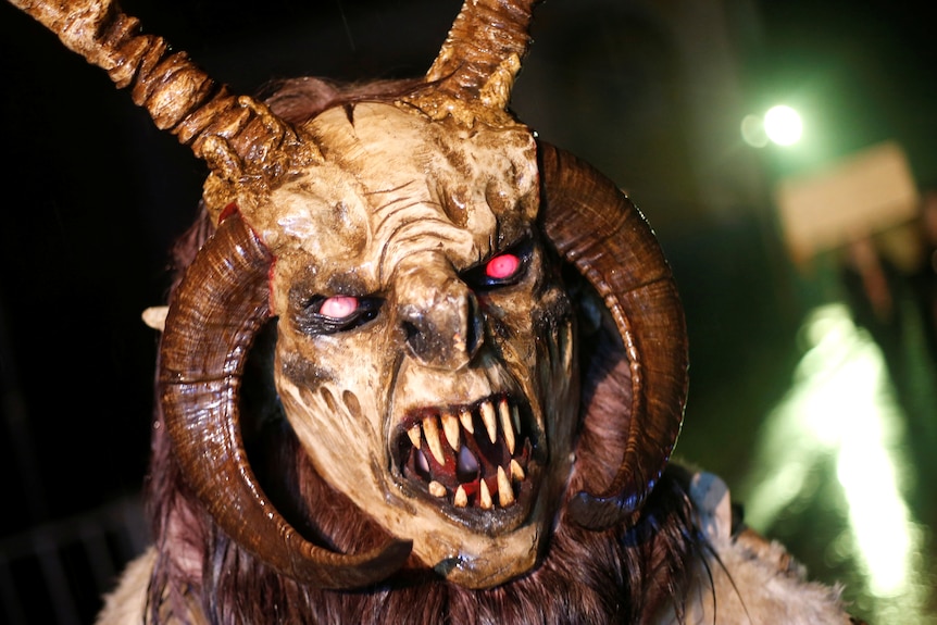 A person wears a demonic goat face mask with horns and sharp large teeth, all carved from wood.