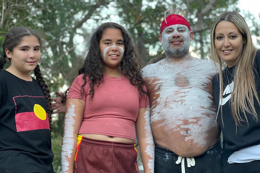 A family - mum, dad and two school-aged daughters - pose for a photo. Dad is in traditional Aboriginal dress