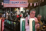 A man wearing a pink shirt and black rimmed glasses pours a beer from a beer tap behind a bar hung with US and British flags