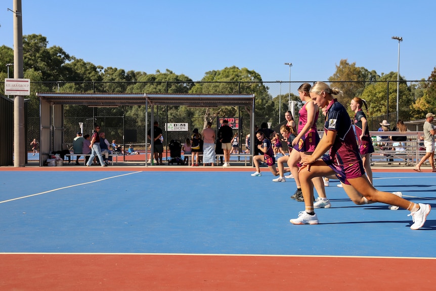 A row of teenage girls stretch on a brightly coloured netball court beneath blue skies