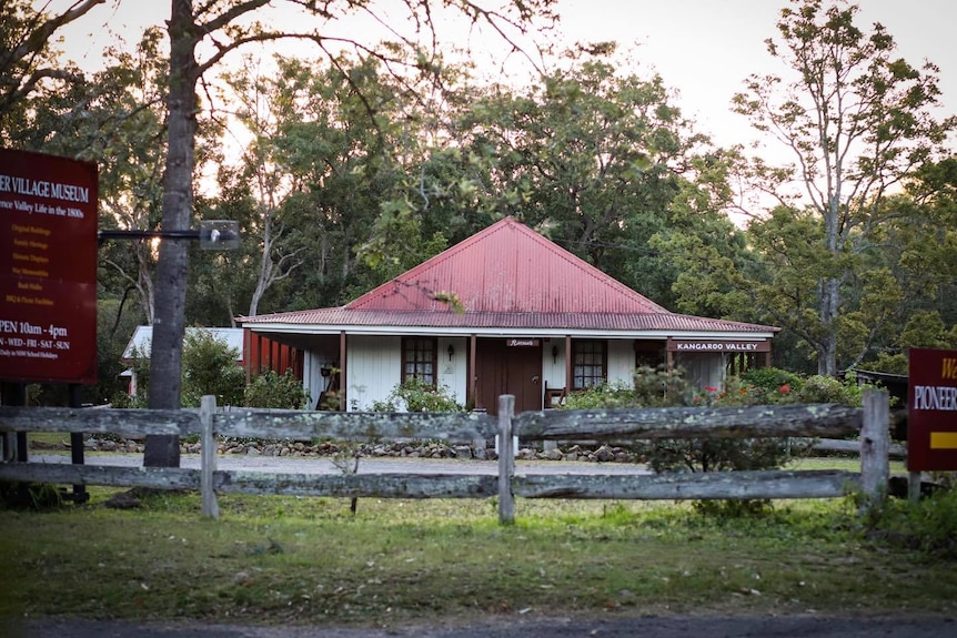 An old house with a red tin roof and white exterior sits on a property behind a wooden fence.