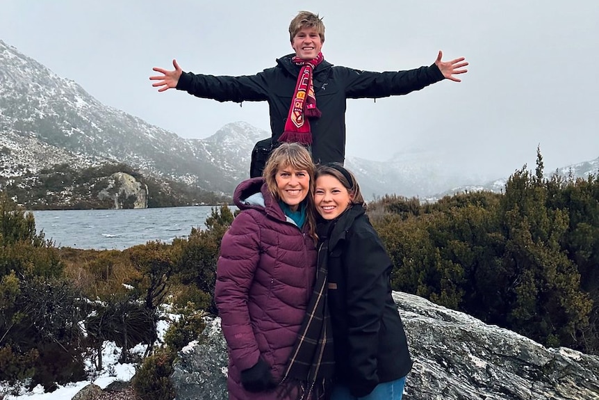 Terri and Bindi Irwin stand with their arms around each other while Robert Irwin stands on a large rock behind them