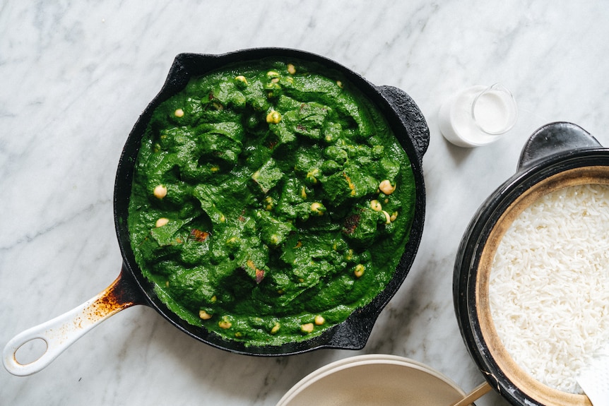 A cast-iron pan filled with spinach and chickpea curry next to a bowl of rice.