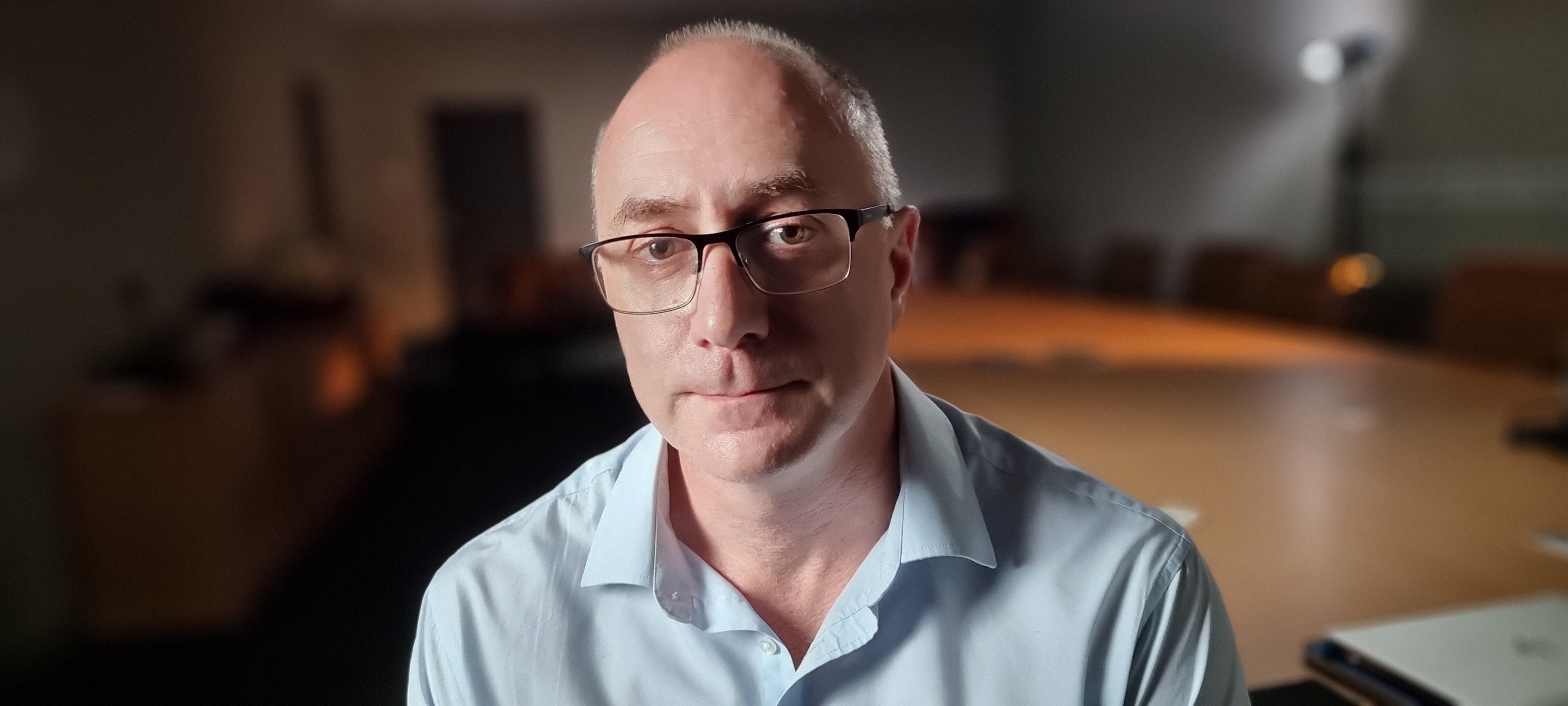 A man wearing glasses sits indoors looking at the camera. Behind him is a large table.