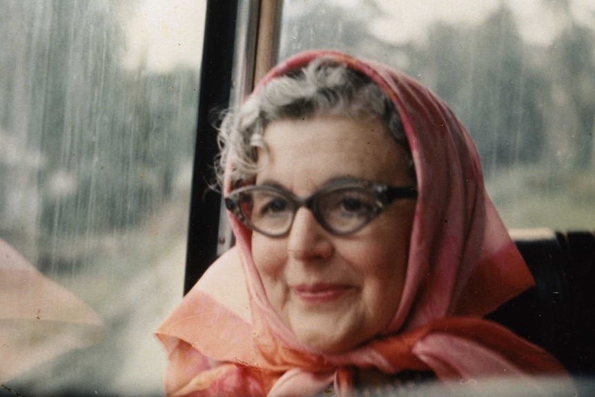A photo of Constance Stokes on a bus, wearing angular glasses and a pink scarf over her head.