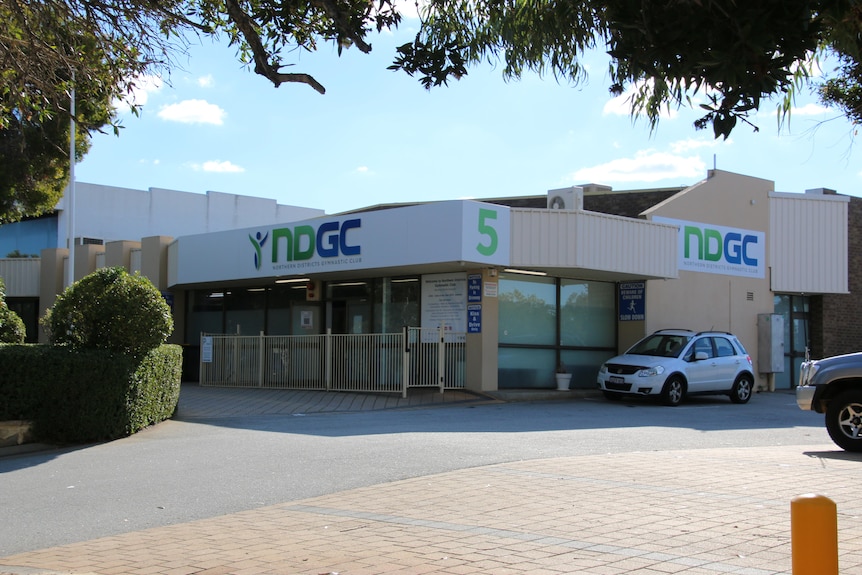 The exterior of a light industrial building with cream brickwork and a white sign on the roof, a parked car and a garden