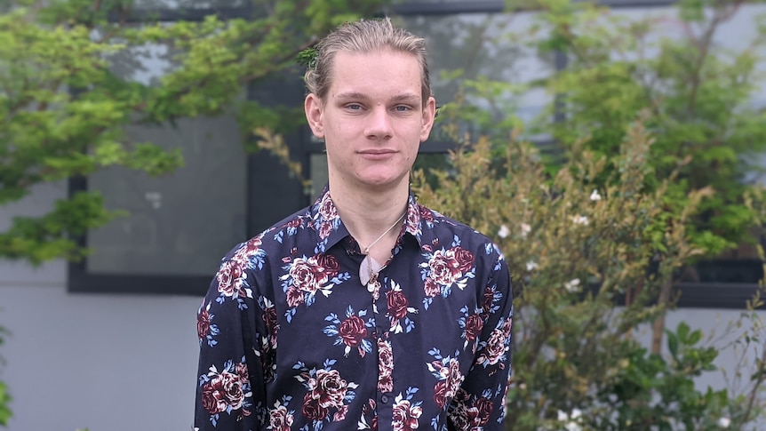 A young man in a flowery shirt stands with his hands behind his back