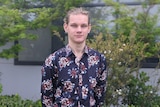 A young man in a flowery shirt stands with his hands behind his back