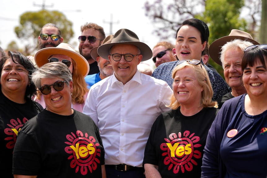 A smiling man in a hat, white shirt and glasses surrounded by smiling people with black shirts saying yes on them