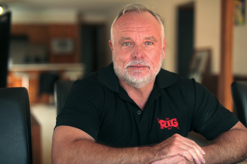 A man sitting inside a house, leans on a table. He has a neutral expression.