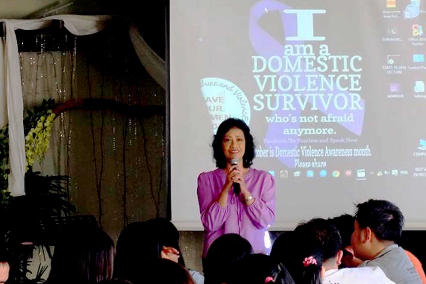 A woman holding a microphone in front of a presentation that reads "I am a domestic violence survivor"