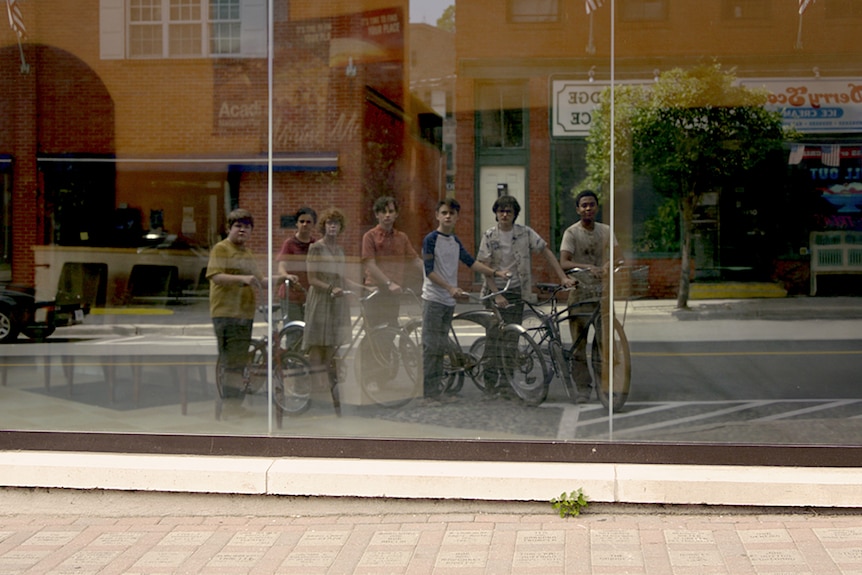 Six male and one female teens stand with their bikes in small town street and look at their reflection on a building exterior.