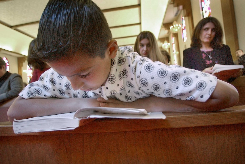 A young boy leans over a Bible as women sit in pews behind him.