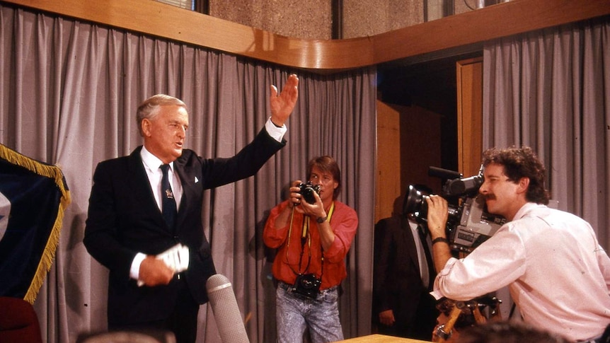 Joh waves his hands from behind a desk as media films.