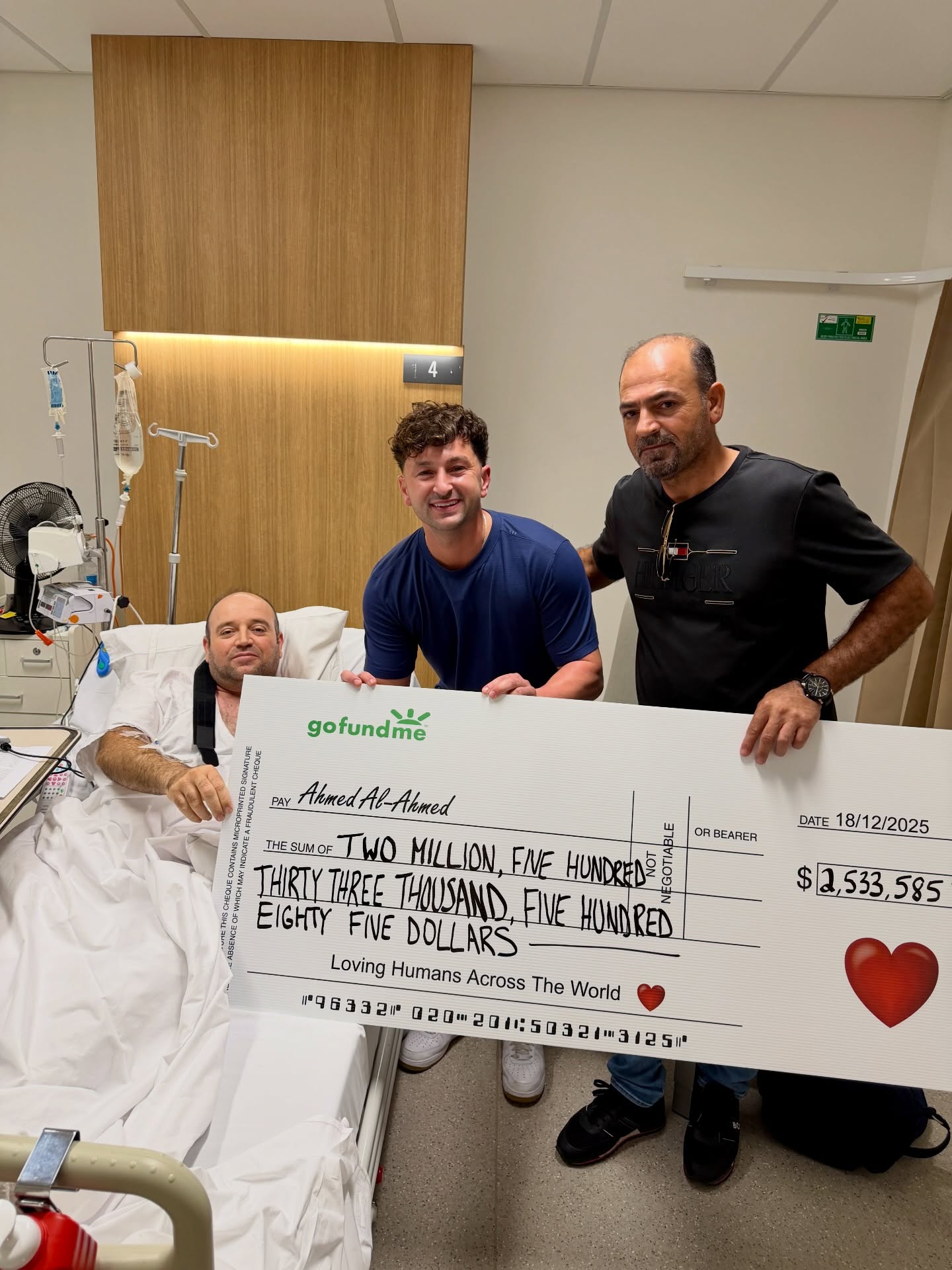 Ahmed Al Ahmed, em uma cama de hospital, posa com outros dois homens segurando um enorme cheque de novidades. 