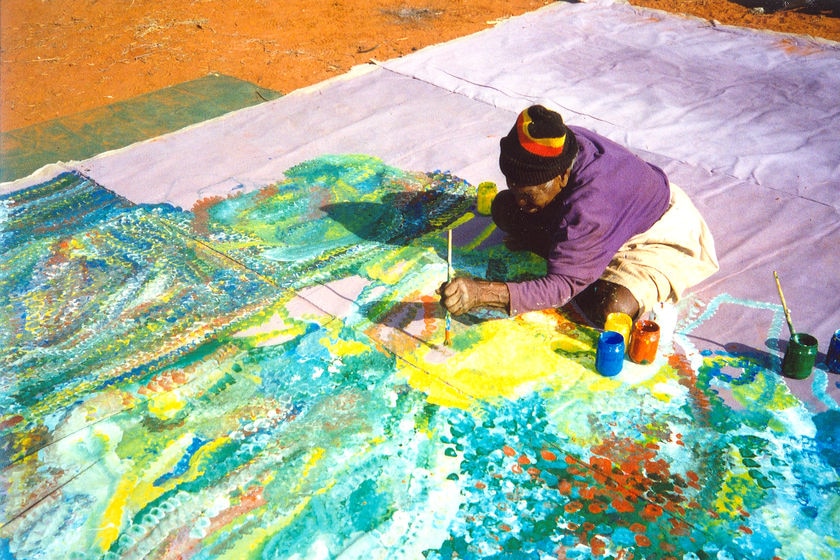 Emily Kngwarreye sitting on a canvas on the ground painting.