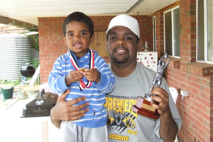 Indigenous man holding a trophy in one hand and his son in the other, who has medal around his neck 