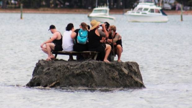 A group of friends sit at a picnic table and drink beer on an island made out of sand away from the shore in coastal waters.