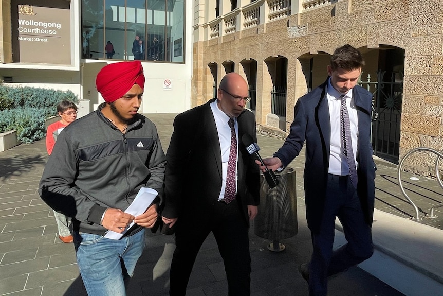 A man in a bright-coloured turban, jacket and jeans, walks alongside two men in dark suits outside a court building.