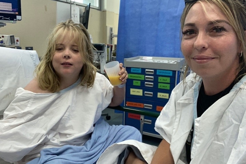 A girl holding a cup of juice, in a hospital bed, wearing white gown, her mum next to her.