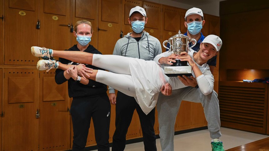 A female tennis player is carried in the arms of three mask-wearing people as she holds a trophy.