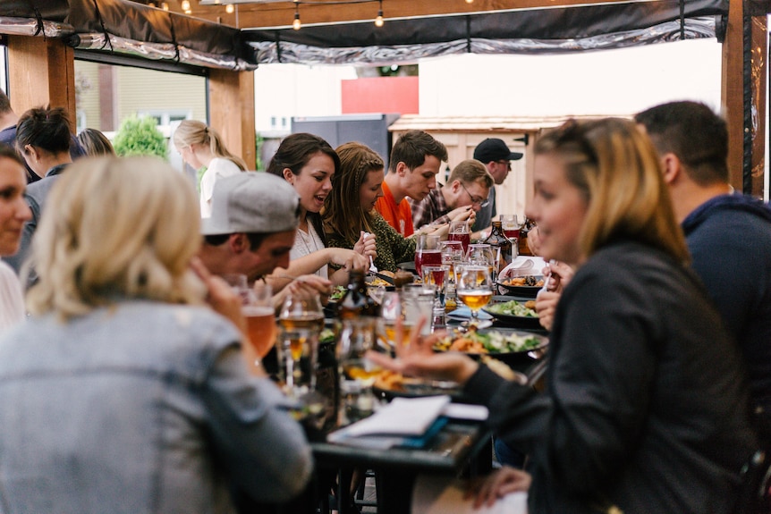 A table of about 15 people chat while eating and drinking wine and beer. 