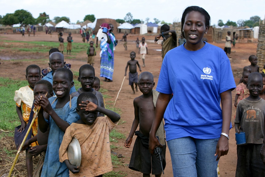 A photo of Dr Barbara Nattabi walking with a group of children with tents in the background.