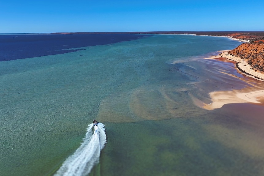 A drone shot of a boat travelling in the water. Beside it is a red cliff. 
