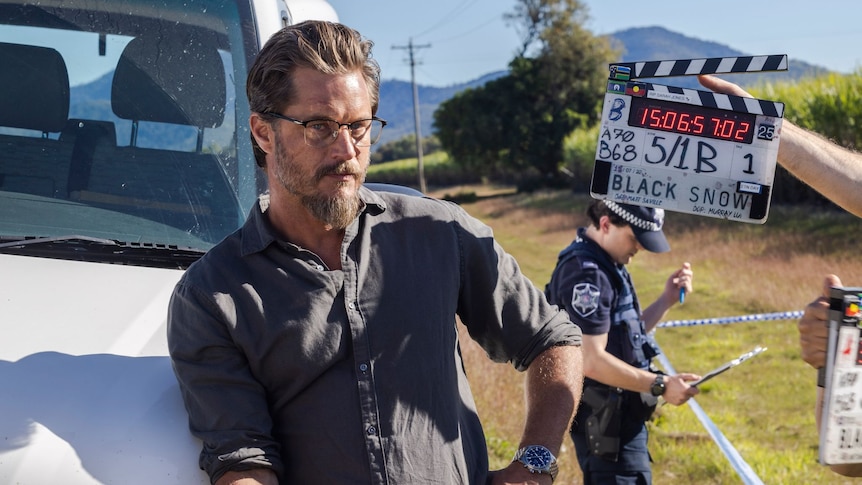 Australian actor Travis Fimmel stands leaning on a car next to a film action sign. 