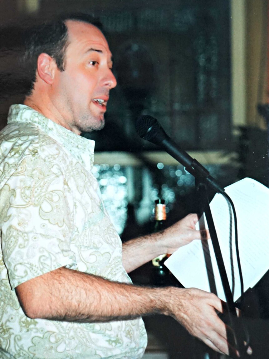A photo of author John Birmingham wearing a paisley shirt and speaking at a microphone in 2002.